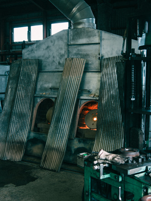 A large industrial forge or furnace with corrugated metal doors, a glowing orange fire inside, and a dark workshop environment with machinery in the foreground.