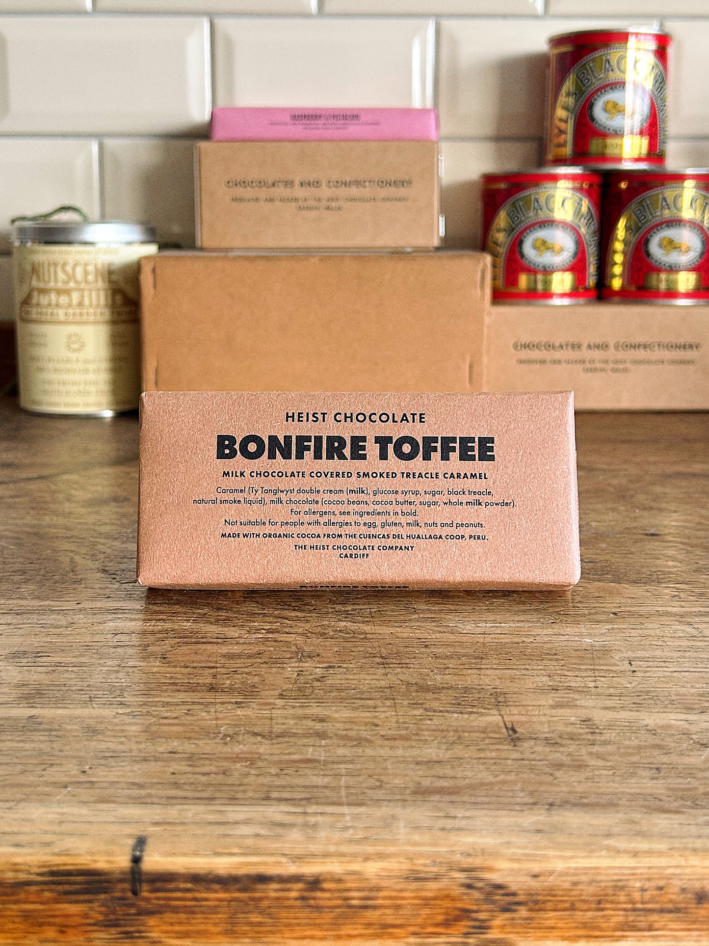 Bar of Heist Chocolate Bonfire Toffee on a wooden surface with other products in the background.