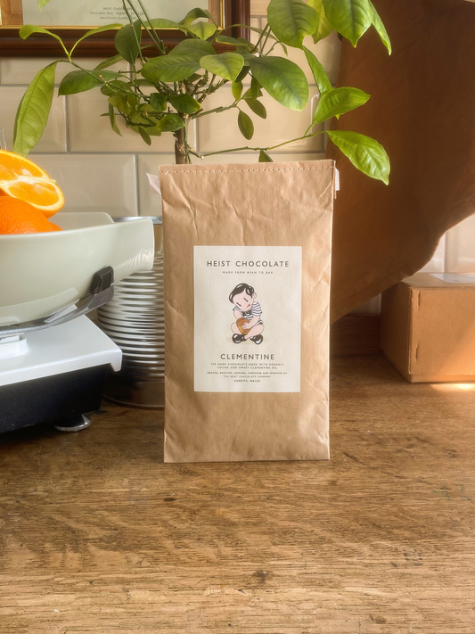 Heist Clementine Chocolate bar in front of a small orange tree on a wooden surface with a tiled background.