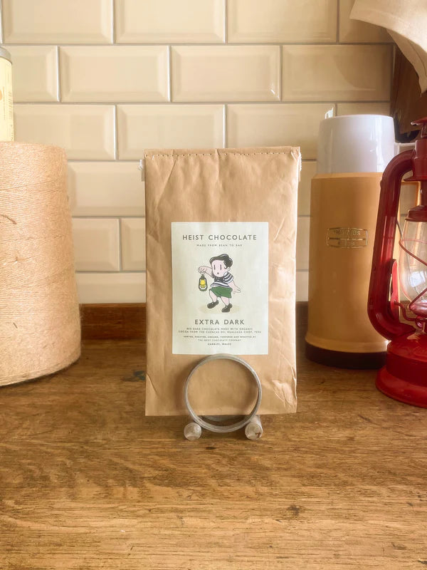 Bar of Heist Extra Dark Chocolate on a wooden surface, with a lantern and thermos in the background.