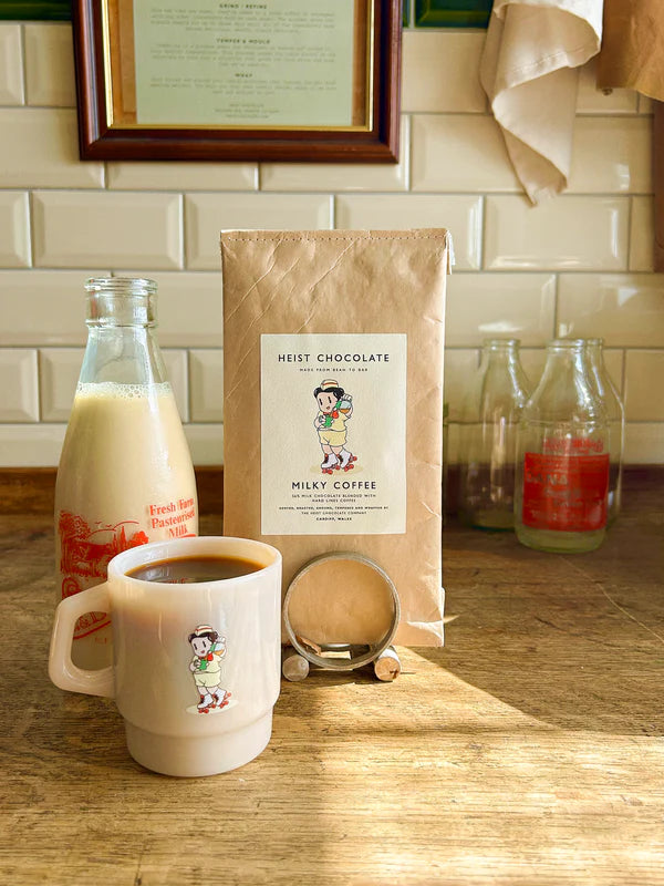 Mug with coffee, bottle of milk, and a bar of bean to bar chocolate, on a wooden surface with a tiled wall background.