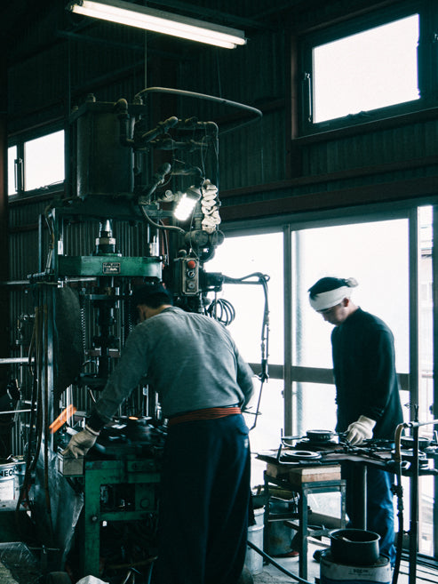 A dark, atmospheric photograph of a traditional blacksmith workshop, showing two men working around a large industrial press machine. One man in a grey shirt and dark trousers is operating the machine, while another man in a black shirt and white headband stands nearby.