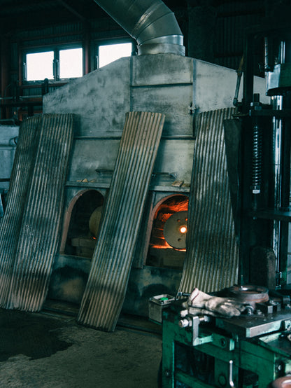 A large industrial forge or furnace with corrugated metal doors, a glowing orange fire inside, and a dark workshop environment with machinery in the foreground.