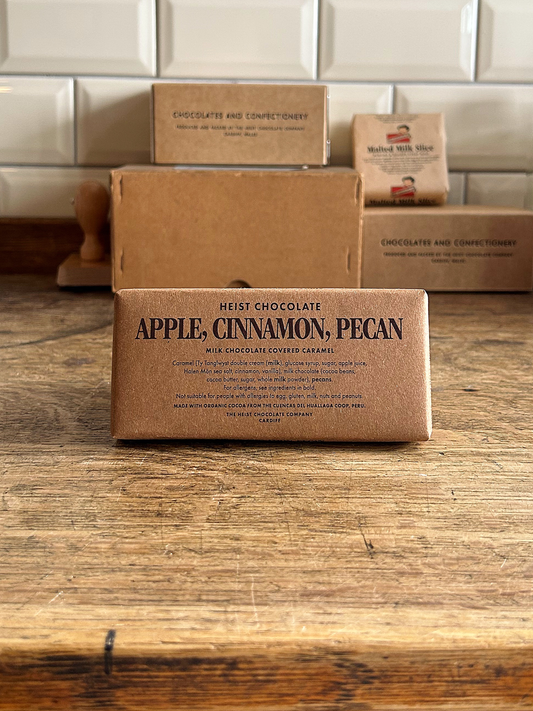 Heist Chocolate Apple, Cinnamon, Pecan bar in brown packaging on a wooden surface with a tiled background.