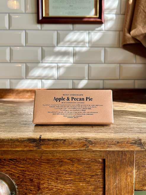 A chocolate bar wrapped in tan paper with 'Apple & Pecan Pie' printed on it, resting on a rustic wooden table in front of a white metro tile wall.