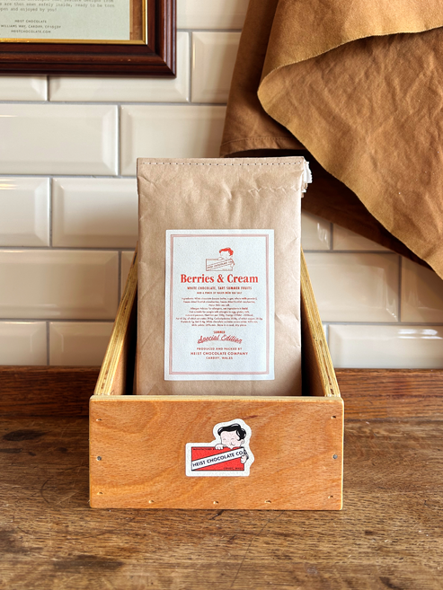 A kraft paper bag labelled 'Berries & Cream' stands in a wooden crate on a rustic wooden table, with a white subway tile wall and brown fabric in the background.