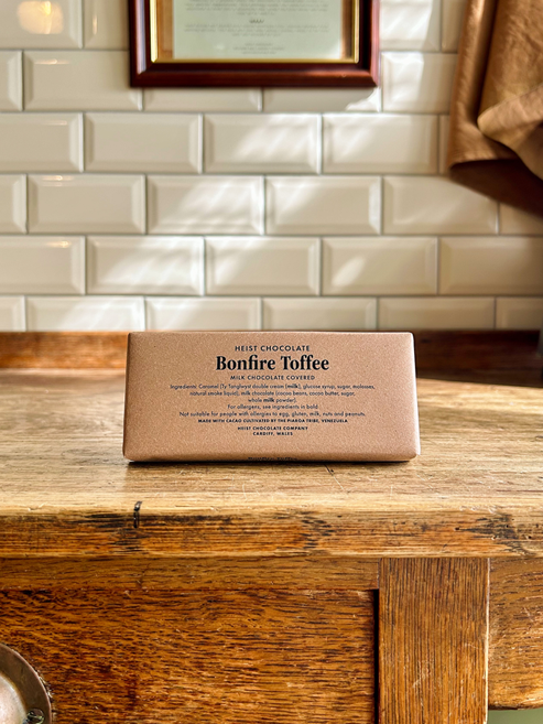 A rectangular bar of Heist Chocolate Bonfire Toffee sits on a rustic wooden table in front of a white metro tile wall.