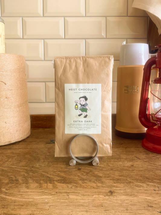 Bar of Heist Extra Dark Chocolate on a wooden surface, with a lantern and thermos in the background. 