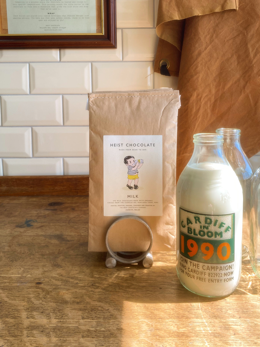 Heist Milk Chocolate bar next to a milk bottle labelled 'Cardiff in Bloom 1990' on a wooden surface with a tiled background.