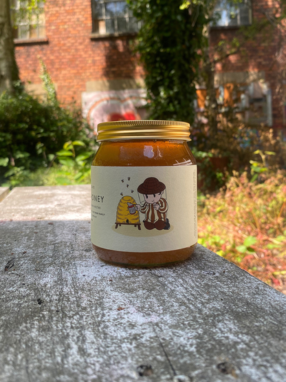 Jar of honey with a depiction of Tompkins tending to a bee hive. The jar is on a wooden surface with a garden background.