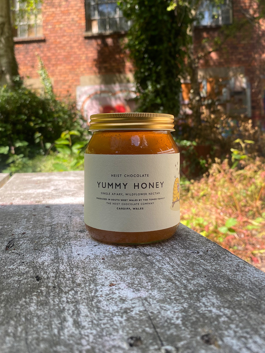 Jar of honey labeled 'Yummy Honey' on a painted, rustic wooden surface with a garden background. A sunny day.