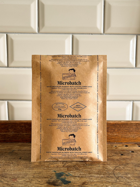 A kraft paper pouch of Microbatch chocolate stands upright on a rustic wooden surface in front of a white metro tile wall.