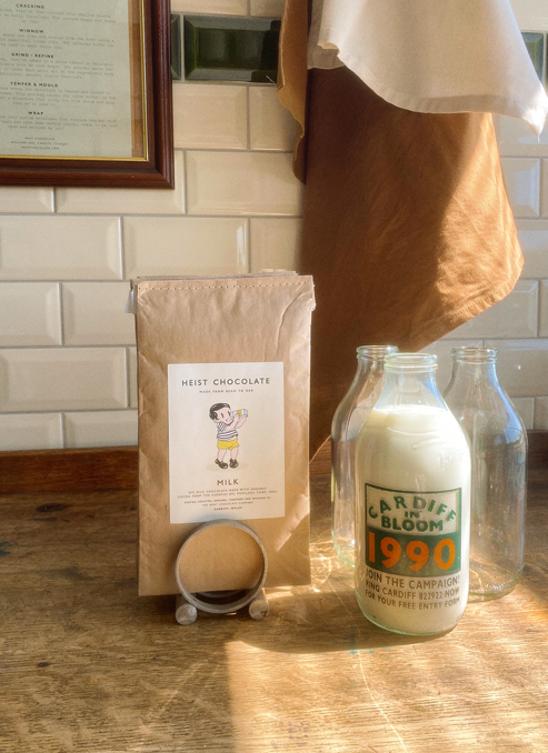 A chocolate bar inside a kraft paper jiffy bag with a stitched top on a wooden desk alongside a full milk bottle with "Cardiff in Bloom, 1990" printed on it.