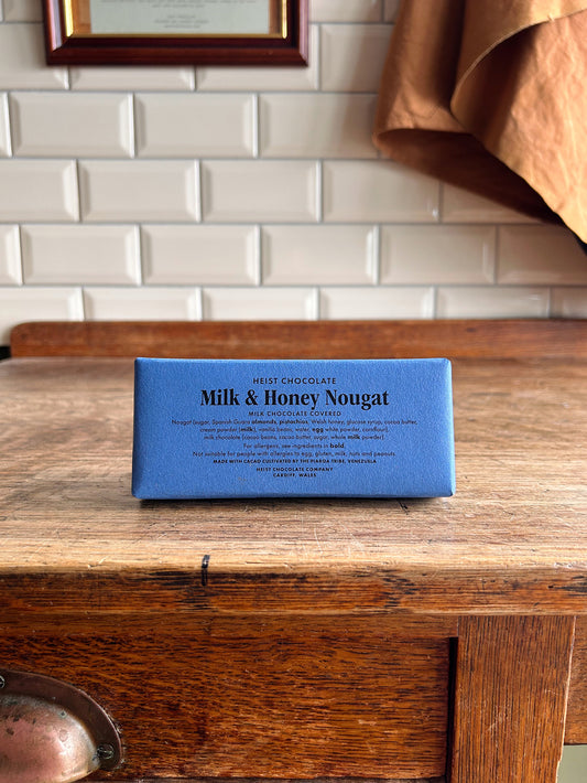 A bean to bar milk chocolate bar in a blue paper wrapper with milk and honey nougat printed on the front in black text.