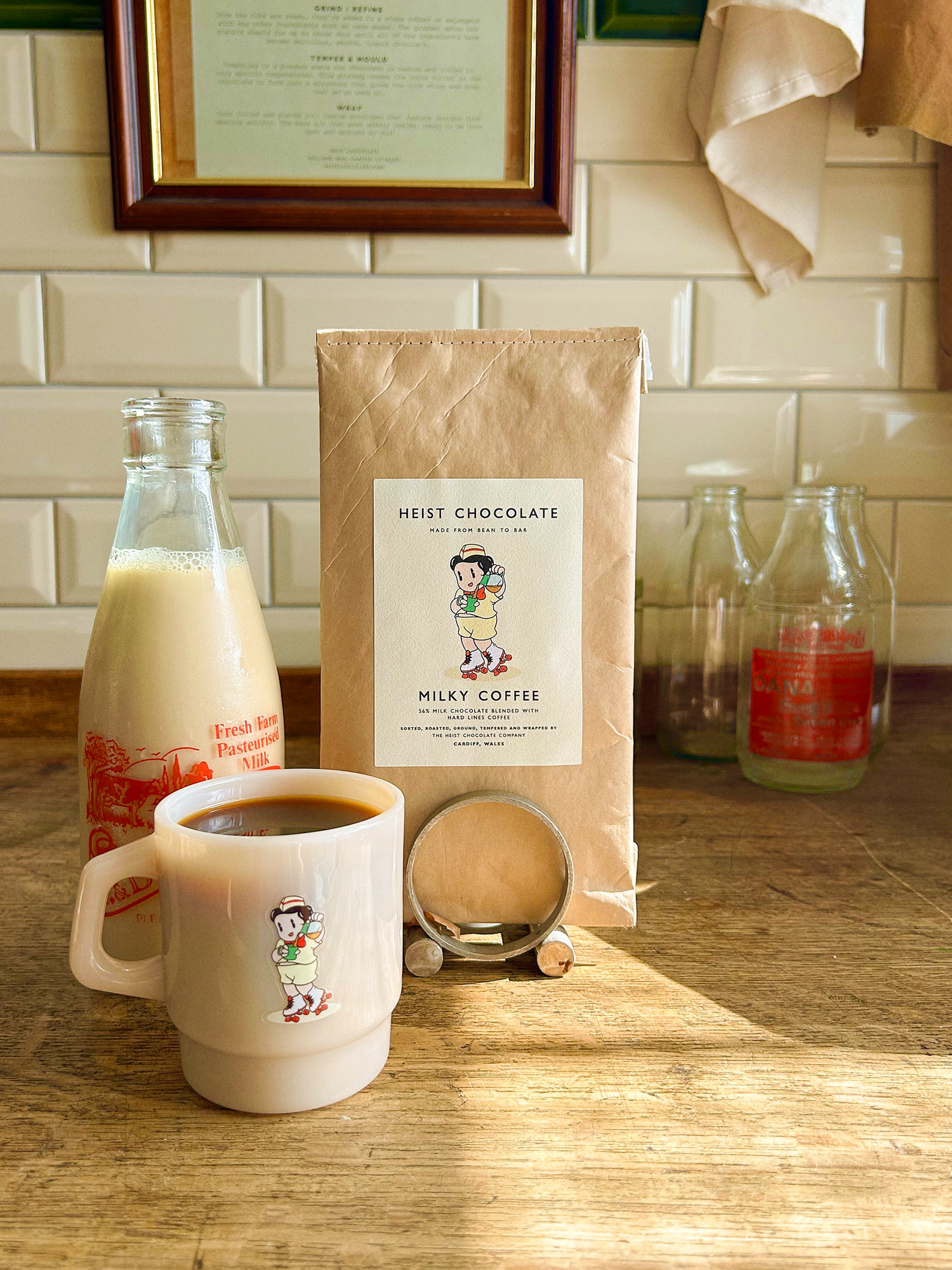 Mug with coffee, bottle of milk, and a bar of bean to bar chocolate, on a wooden surface with a tiled wall background.