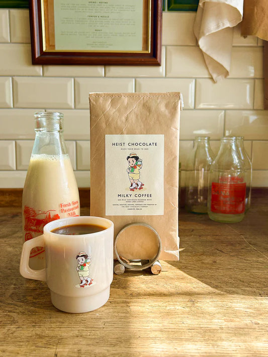 Mug with coffee, bottle of milk, and a bar of bean to bar chocolate, on a wooden surface with a tiled wall background.