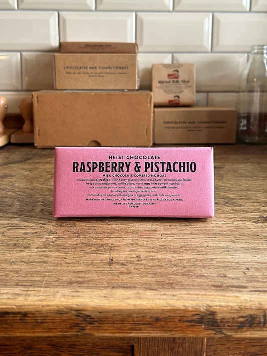 Bean to bar chocolate and nougat bar in pink paper packaging with black text on the front on a wooden surface with a tiled background.