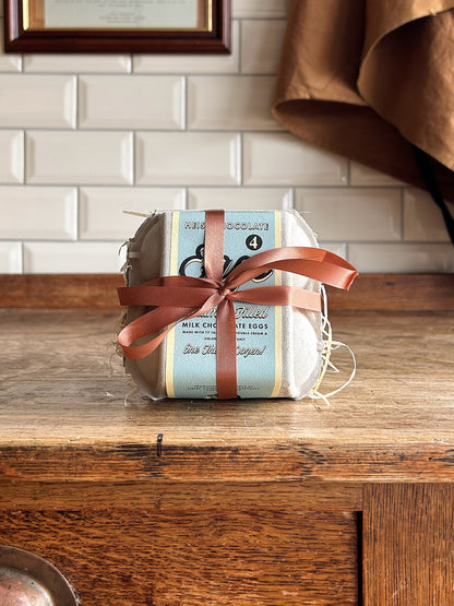 A box of 4 Easter eggs with a blue label and brown ribbon. Sat on a wooden table with a tiled background.
