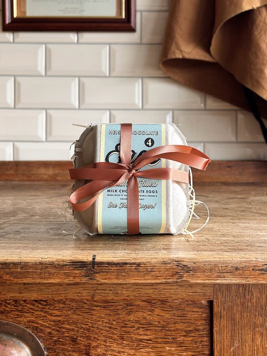 A box of 4 Easter eggs with a blue label and brown ribbon. Sat on a wooden table with a tiled background.