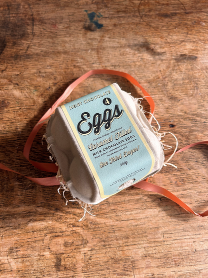 A box of 4 Easter eggs with a blue label and brown ribbon. Sat on a wooden table with a tiled background.