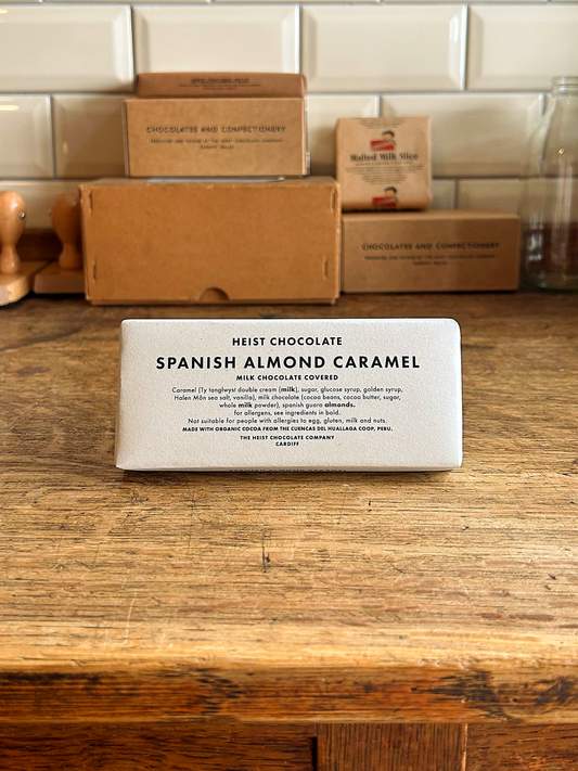 Bar of Heist Chocolate Spanish Almond Caramel on a wooden surface with packaging in the background.