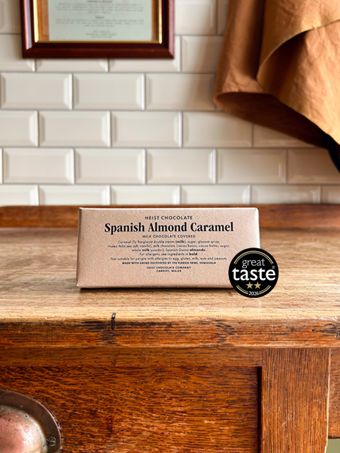 A wrapped bar of chocolate labelled 'Spanish Almond Caramel' sits on a rustic wooden table in front of a cream metro tile wall. A round 'great taste 2026' award badge is placed next to the package.
