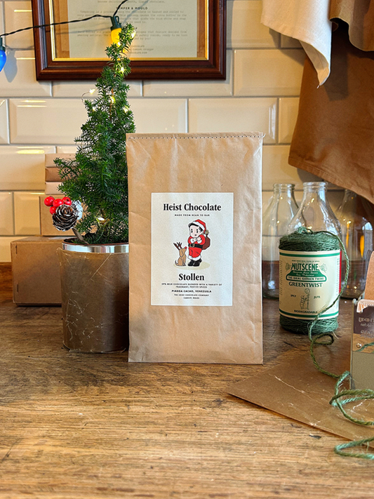 Heist Chocolate Stollen package on a wooden surface with decorative items in the background, including a small Christmas tree and a reel of twine.
