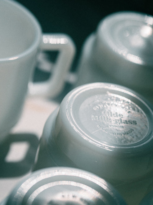 A close-up, artistic shot of several vintage white ceramic mugs and cups, with one cup lying on its side showing the embossed brand name 'Olde Milk Glass, Japan' on the bottom.