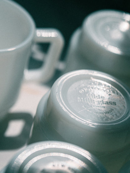 A close-up, artistic shot of several vintage white ceramic mugs and cups, with one cup lying on its side showing the embossed brand name 'Olde Milk Glass, Japan' on the bottom.