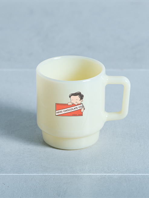 A pale cream-colored ceramic mug with a handle, featuring a cartoon character and the text 'HEIST CHOCOLATE CO' printed on the front, set against a light grEy background.