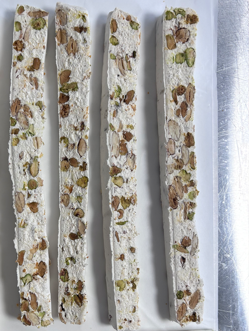 Four long, narrow strips of nougat filled with almonds and pistachios, arranged vertically on a white rectangular plate next to a stainless steel surface.